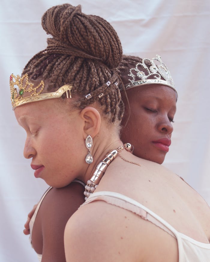 Close-up of two African women with braided hair and tiaras, embracing and radiating elegance.