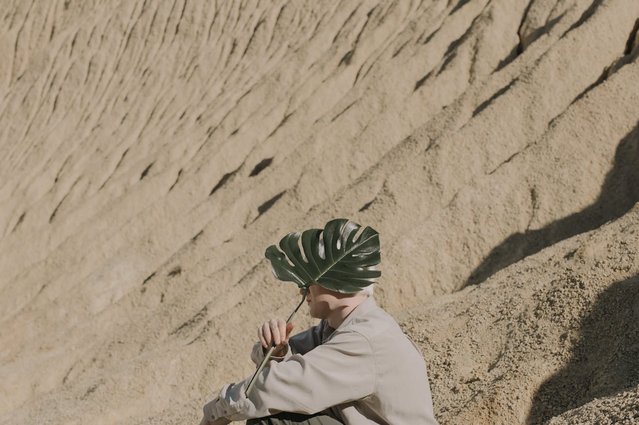 A person shields their face with a leaf in a sandy desert environment, creating a unique and intriguing visual.